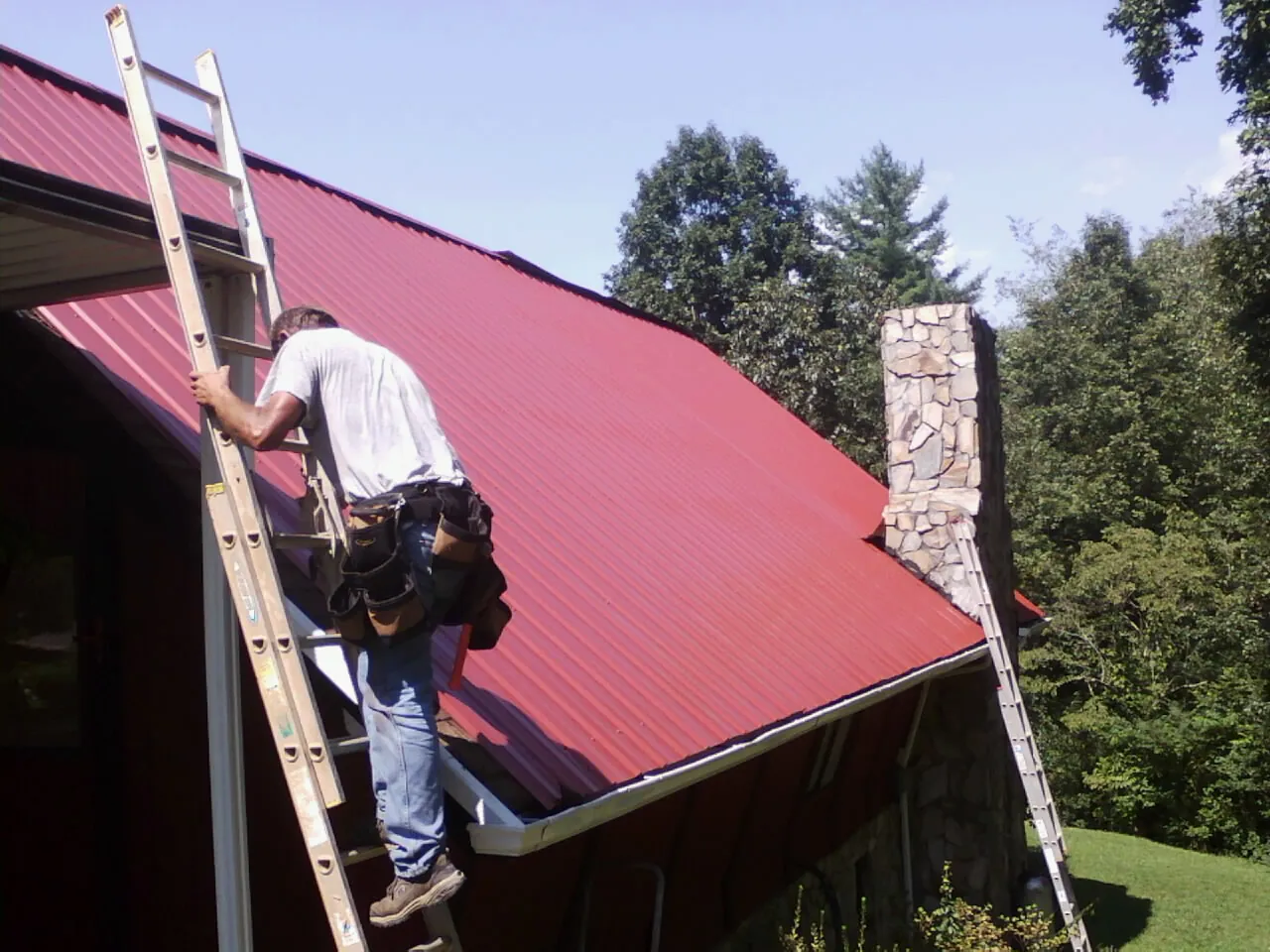 Roofer on a ladder beside a red metal roof and stone chimney — Soffit & Fascia Repair by Roofing Brothers in Jasonville
